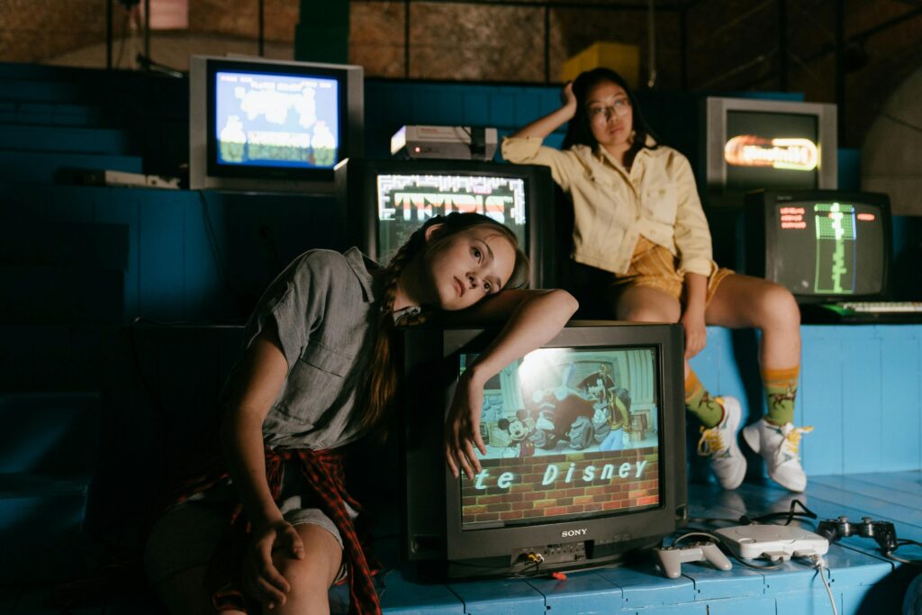 Two women in casual outfits immersed in retro gaming with vintage televisions and game consoles.